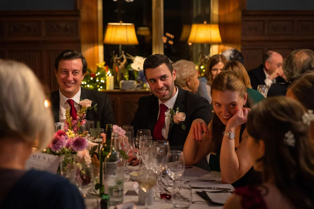 Wedding guests laugh and smile around a candlelit banquet table during the wedding breakfast speeches in a wood-panelled dining room.