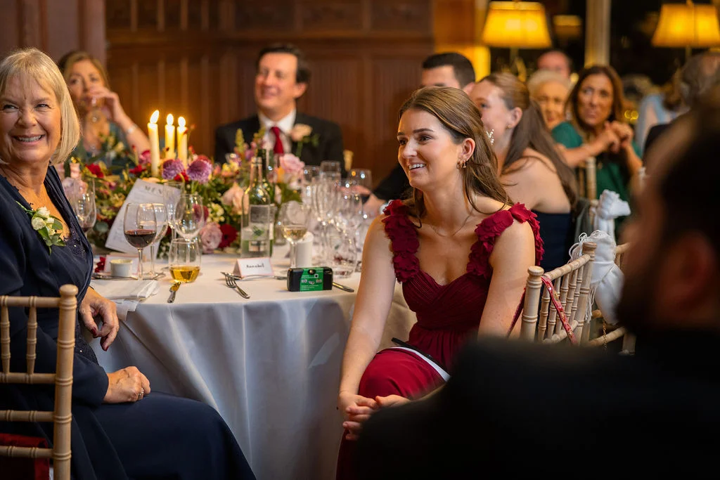 Smiling guests seated at a round dining table during wedding speeches in a wood-panelled reception room.