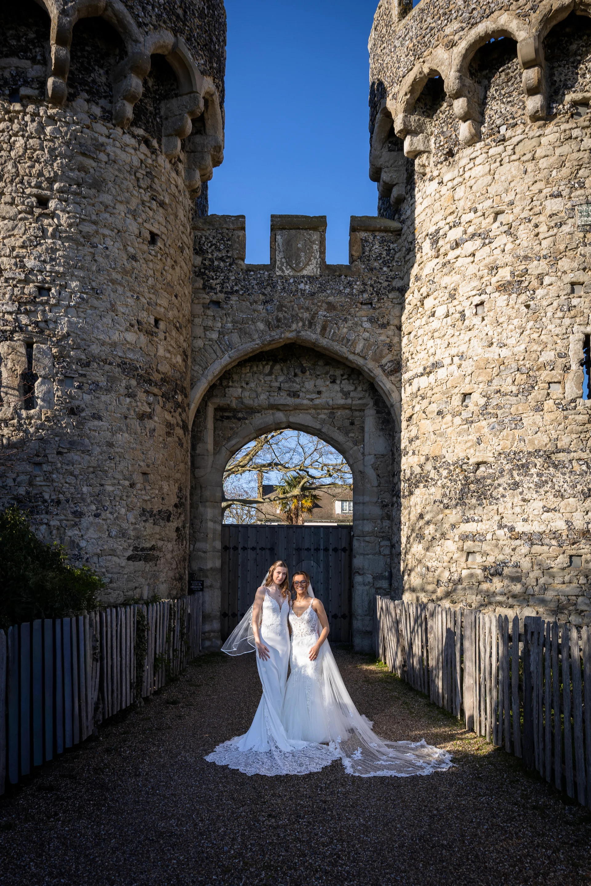Two brides pose proudly together in their wedding gowns framed by the magnificent stone towers and arched gateway of a historic castle.