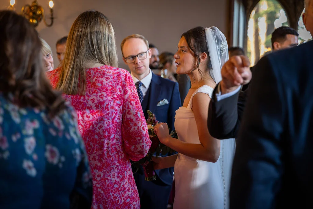 A smiling bride wearing a simple white column dress and veil chats animatedly with wedding guests during a relaxed drinks reception.