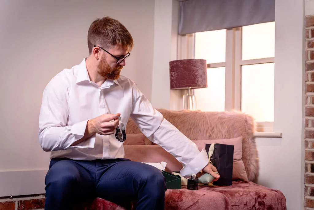 A groom sitting on a pink sofa in a white shirt and trousers unwraps a gift and examines a watch during morning preparations.