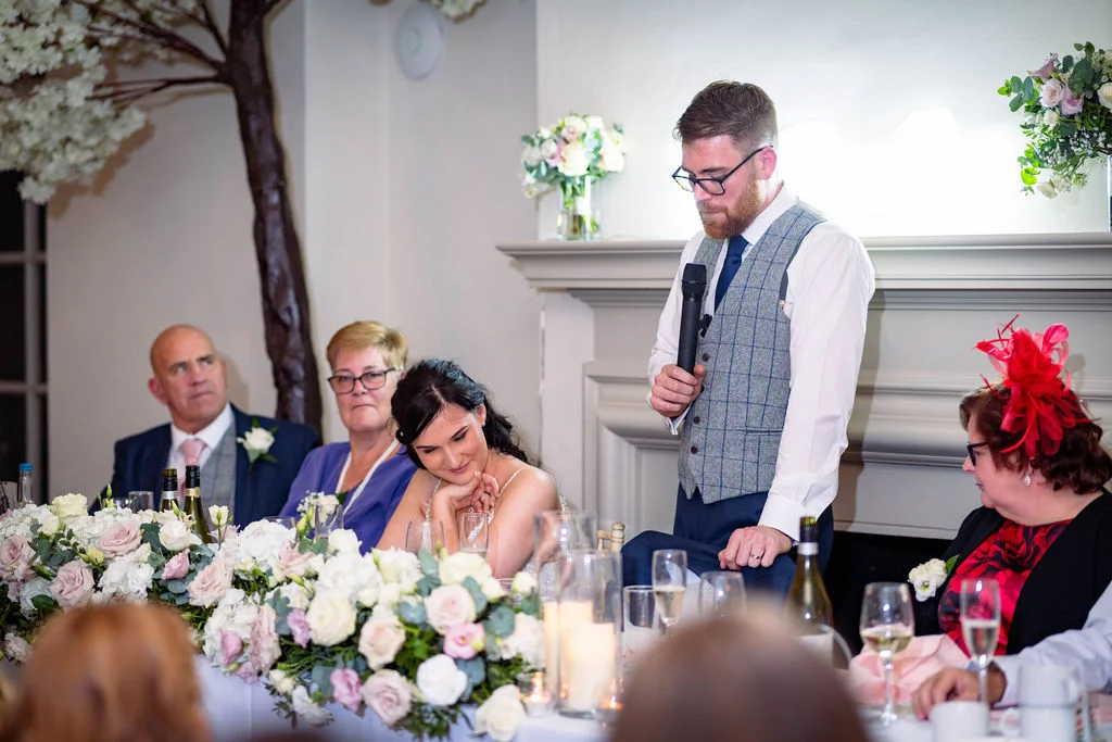 A groom in a tweed waistcoat delivers a speech at the top table while the bride looks on tenderly with family seated beside them.