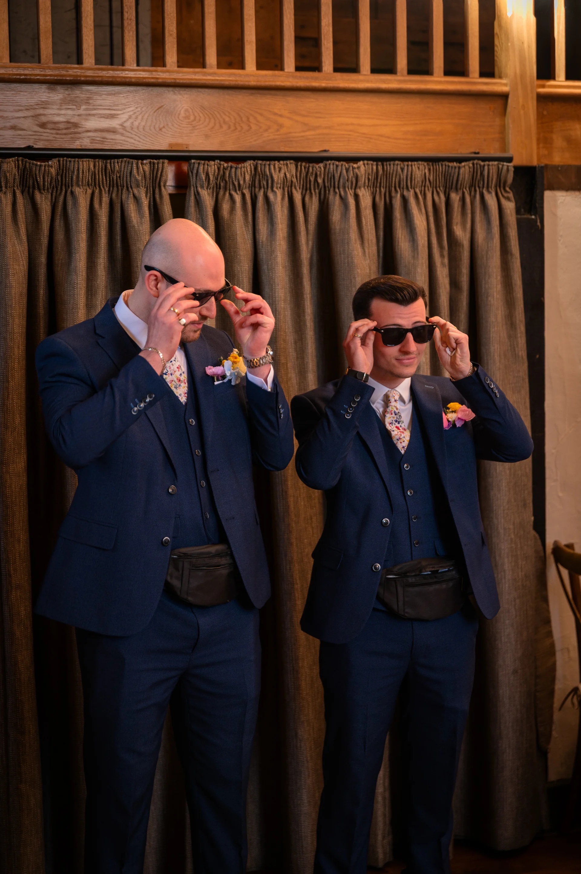 Two grooms in matching navy three-piece suits adjust their sunglasses while posing indoors in front of draped curtains.