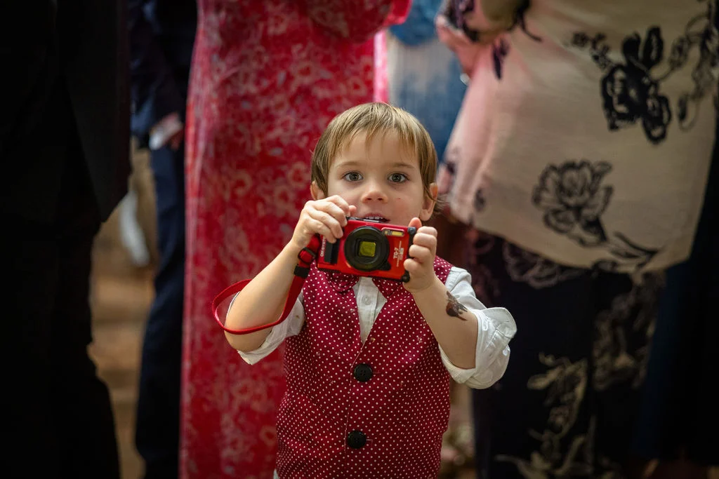 A young page boy wearing a red polka-dot waistcoat holds up a small red camera to take a photo amidst wedding guests.