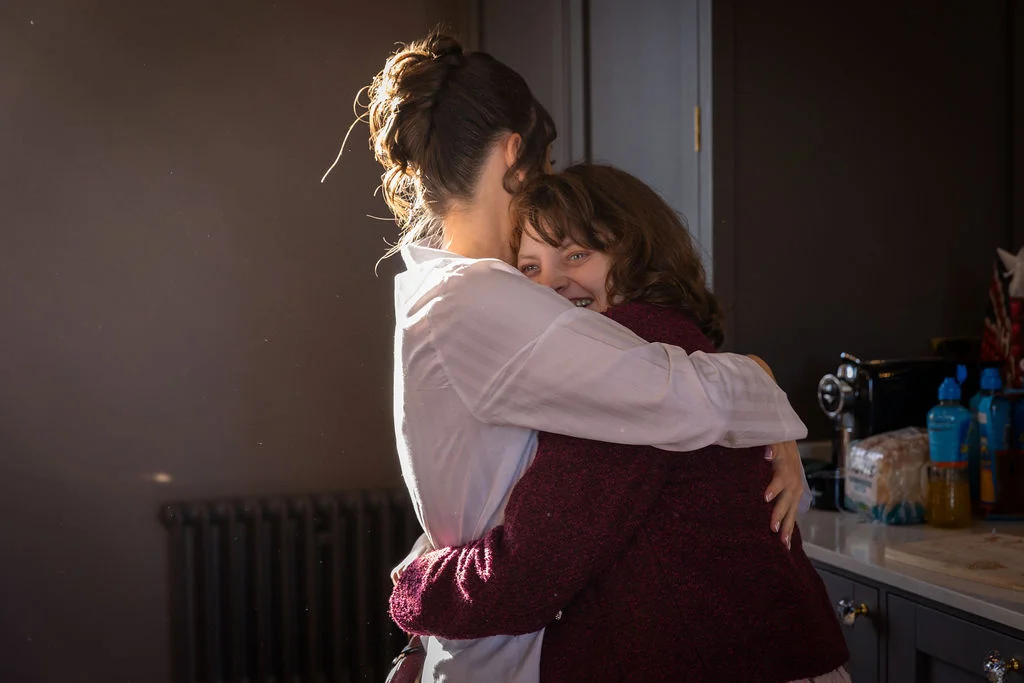 A tender, emotional embrace shared between two people in a warmly lit room during morning preparations.