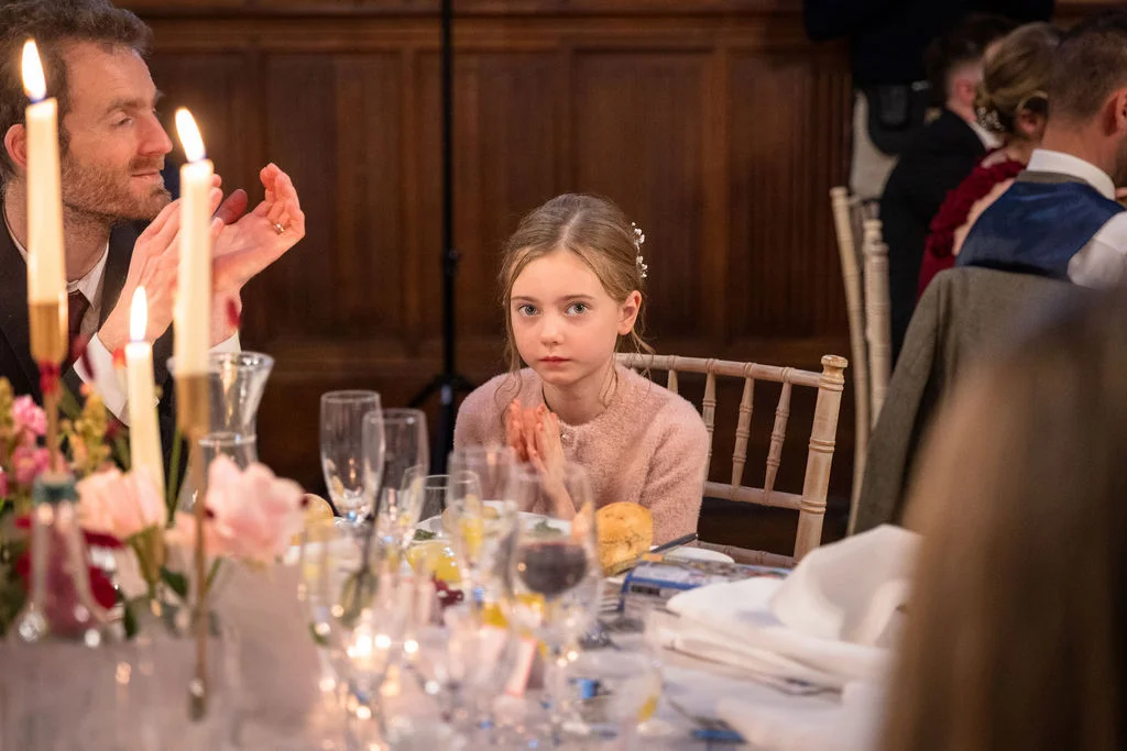 A young flower girl sits attentively at a wedding breakfast table during speeches, with guests clapping softly in the warmly lit room.
