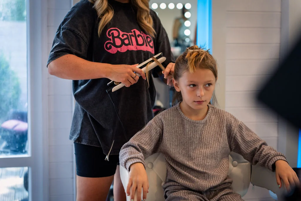 A young girl sits patiently in a white styling chair while having her hair straightened during wedding morning preparations.