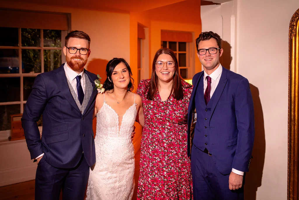 A smiling bride in a lace gown and groom in a navy suit pose alongside two guests indoors under warm lighting.
