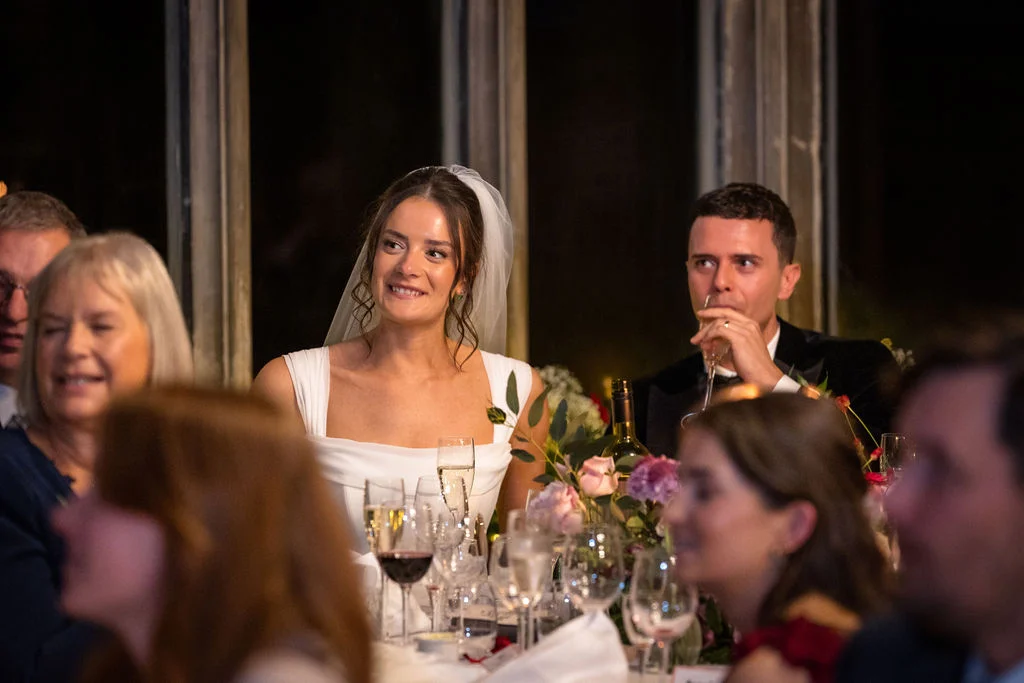 The bride smiles warmly while seated at the top table during wedding speeches, with the groom drinking champagne beside her and guests surrounding them.