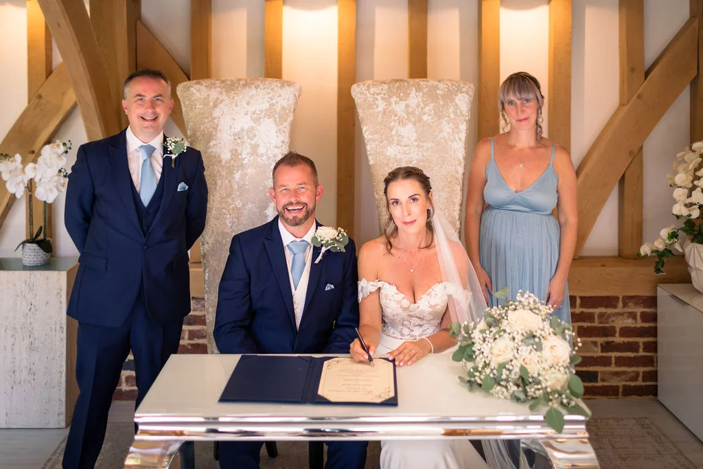 The bride and groom smile while signing the marriage register at a barn wedding, flanked by a groomsman in a navy suit and a bridesmaid in a blue dress.