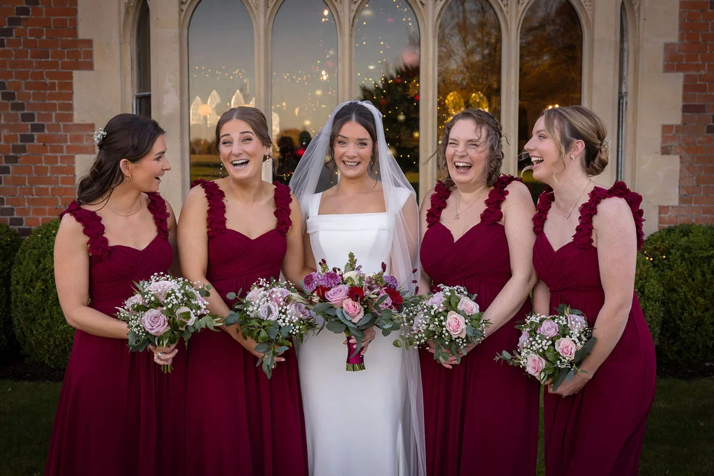 A smiling bride in a white gown and veil stands at the centre of her four bridesmaids, who are all wearing matching burgundy dresses and laughing outside a historic stone building.