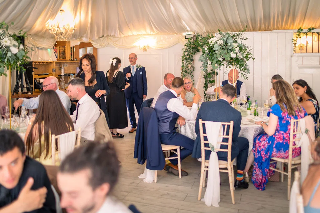 Wedding guests mingle and listen to speeches inside a brightly lit marquee featuring draped ceilings and elegant floral arrangements.