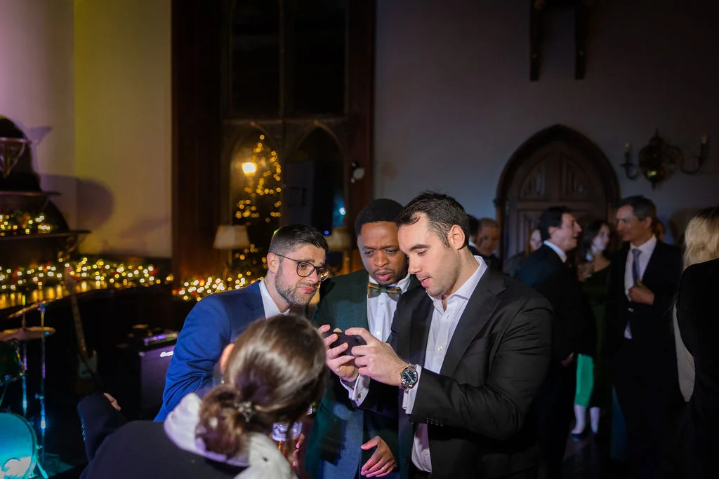 A group of wedding guests gather around a smartphone at an evening reception, illuminated by warm ambient lighting inside an atmospheric venue.
