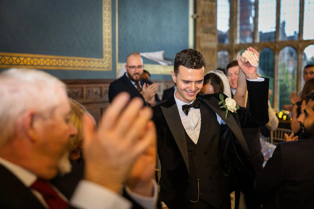A smiling groom in a black tuxedo and waistcoat walks into the reception space holding hands up high, surrounded by cheering guests.