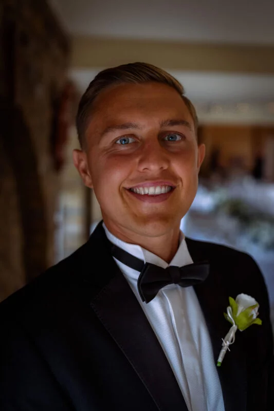 A smiling groom wearing a classic black tuxedo, white shirt, black bow tie, and a white rose buttonhole.