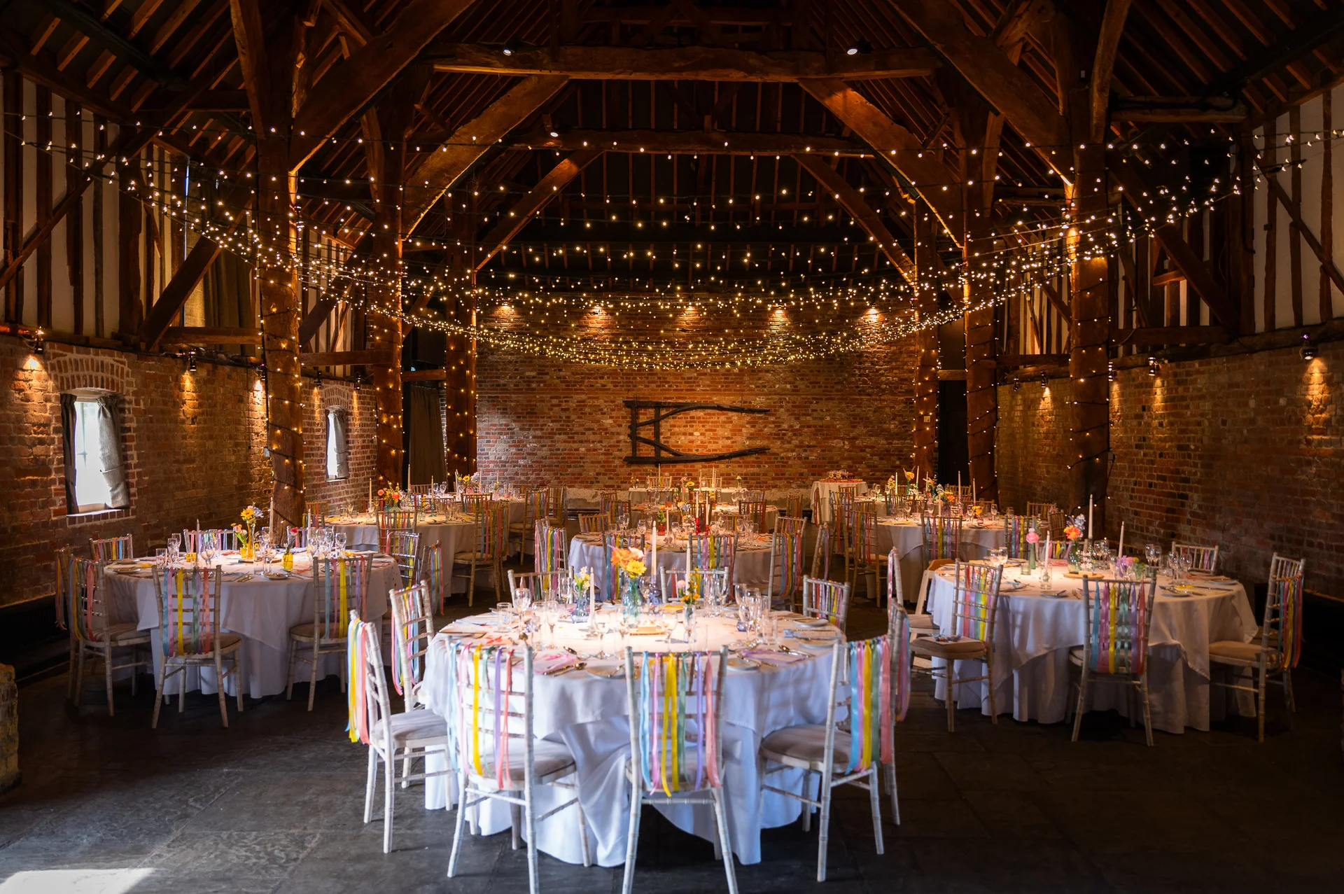 An atmospheric shot of a rustic brick and oak framed barn wedding reception venue featuring round tables decorated with white linens and colourful ribbons tied to the chairs beneath a magnificent fairy light canopy.