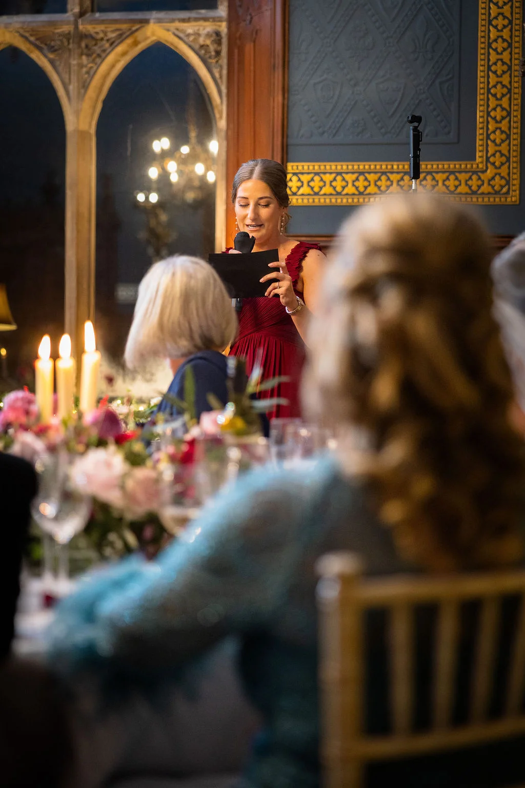 A bridesmaid in a burgundy dress delivers a heartfelt speech during the wedding reception, illuminated by candlelight near large arched windows.