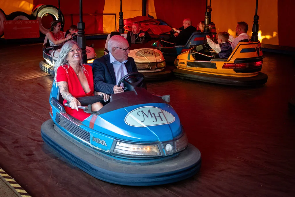 Wedding guests enjoy riding bumper cars together inside a colourful funfair tent during an evening reception celebration.