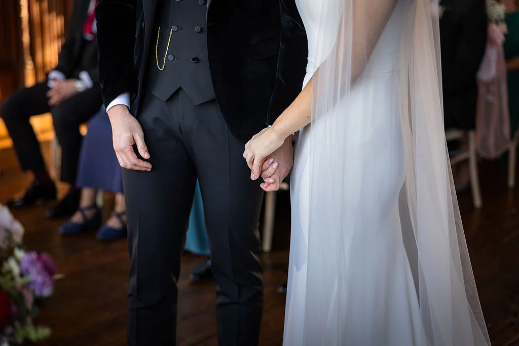 Close-up shot of a newlywed couple holding hands during their ceremony, featuring the groom in a black suit with a velvet jacket and waistcoat, and the bride in a sleek white crepe gown with a sheer veil.