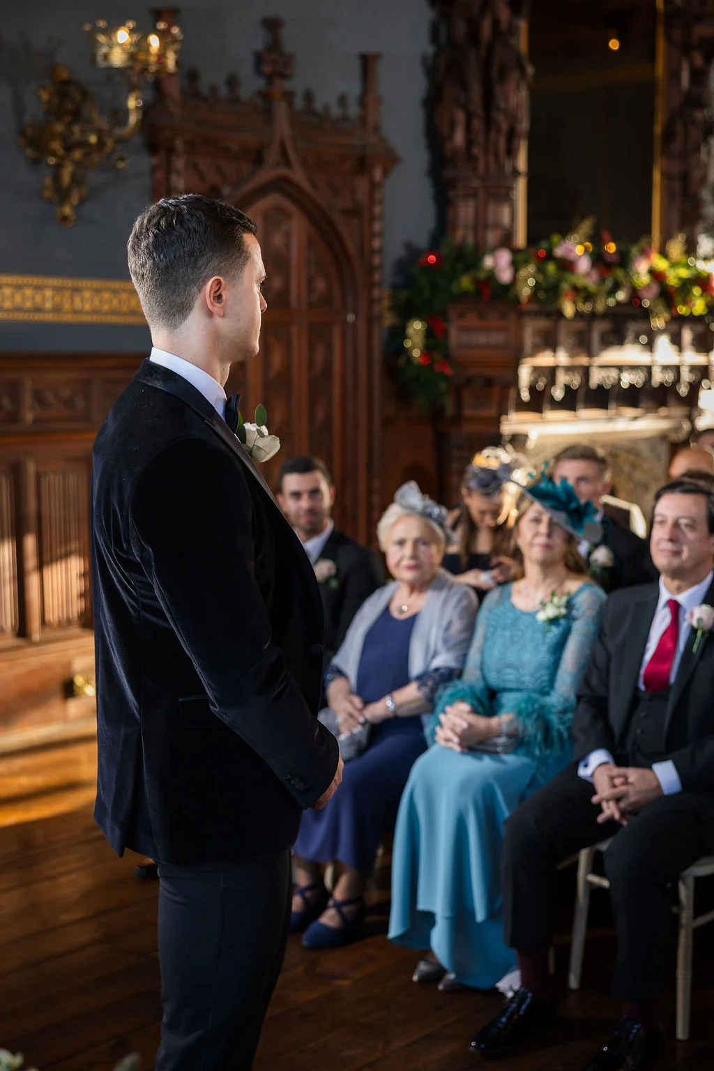 An over-the-shoulder shot captures a groom in a black velvet tuxedo waiting at the altar inside a wood-panelled stately home while seated guests watch.