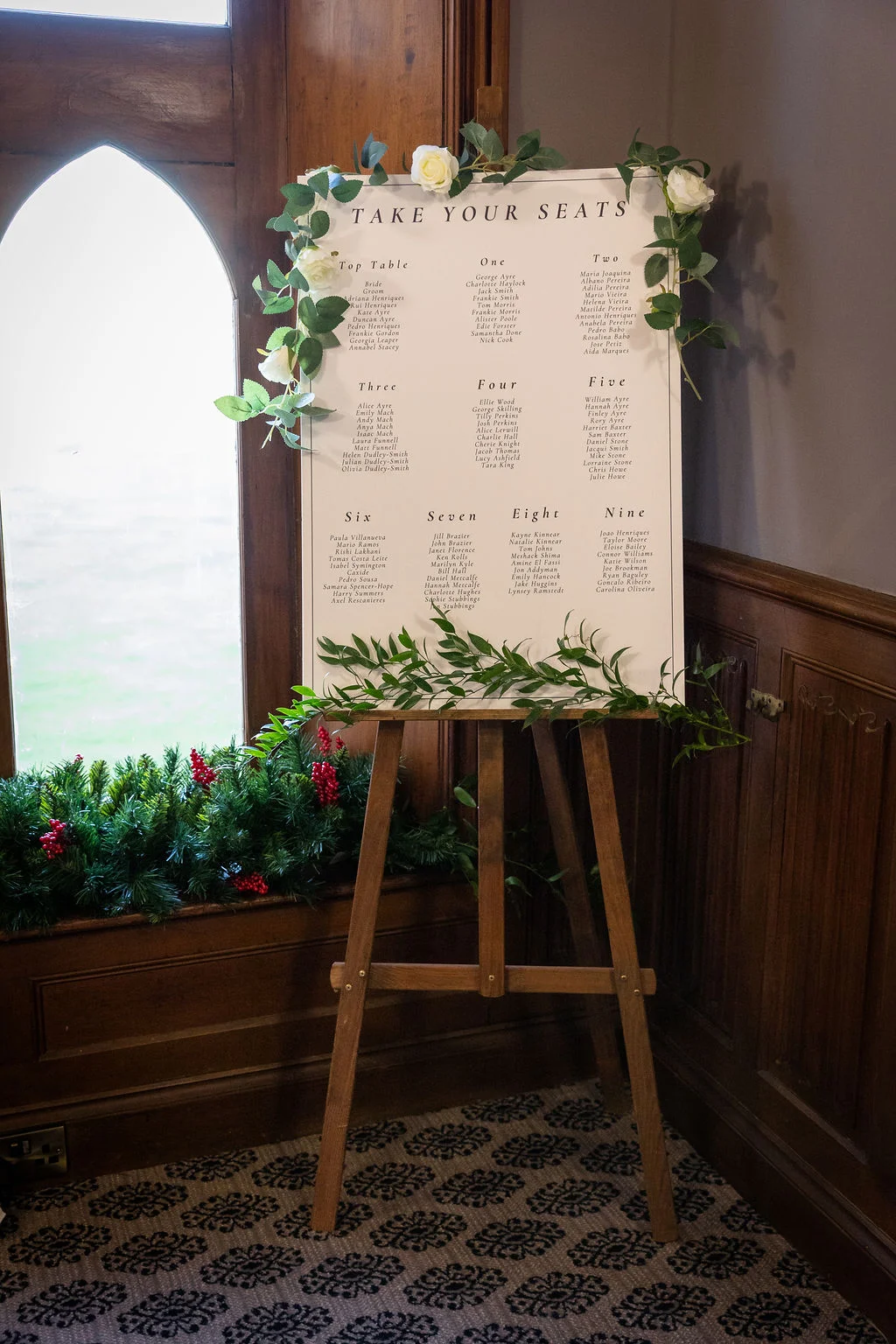 A white wedding seating plan titled 'Take Your Seats' is displayed on a wooden easel, decorated with white roses and trailing greenery.