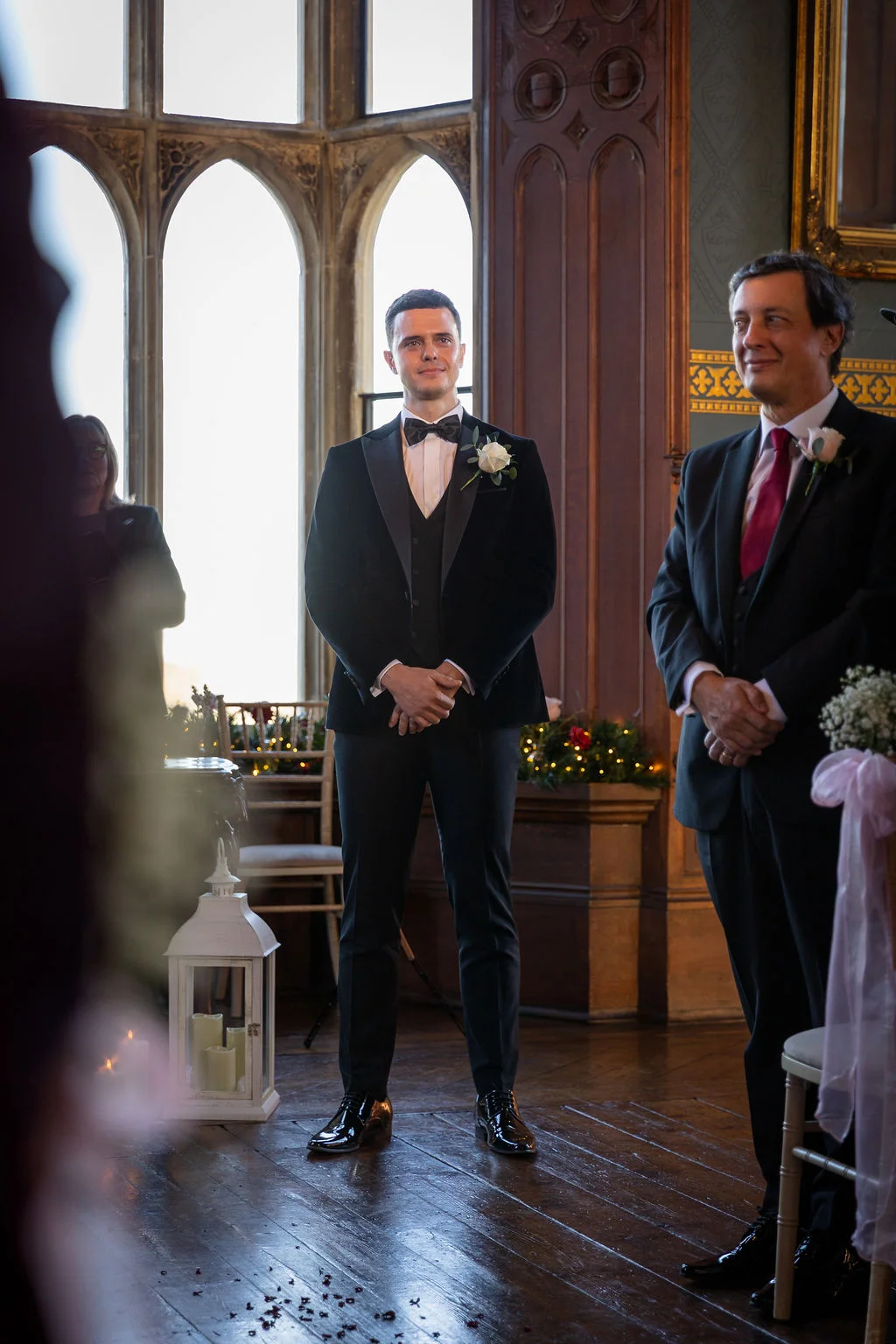 A handsome groom in a velvet tuxedo waits attentively at the altar alongside his best man inside a grand historic venue with tall arched windows.
