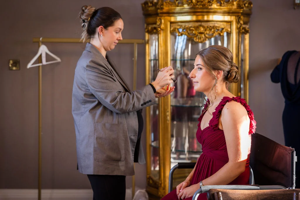 A bridesmaid having her makeup applied and hair styled in a low bun while wearing a ruffled burgundy dress.