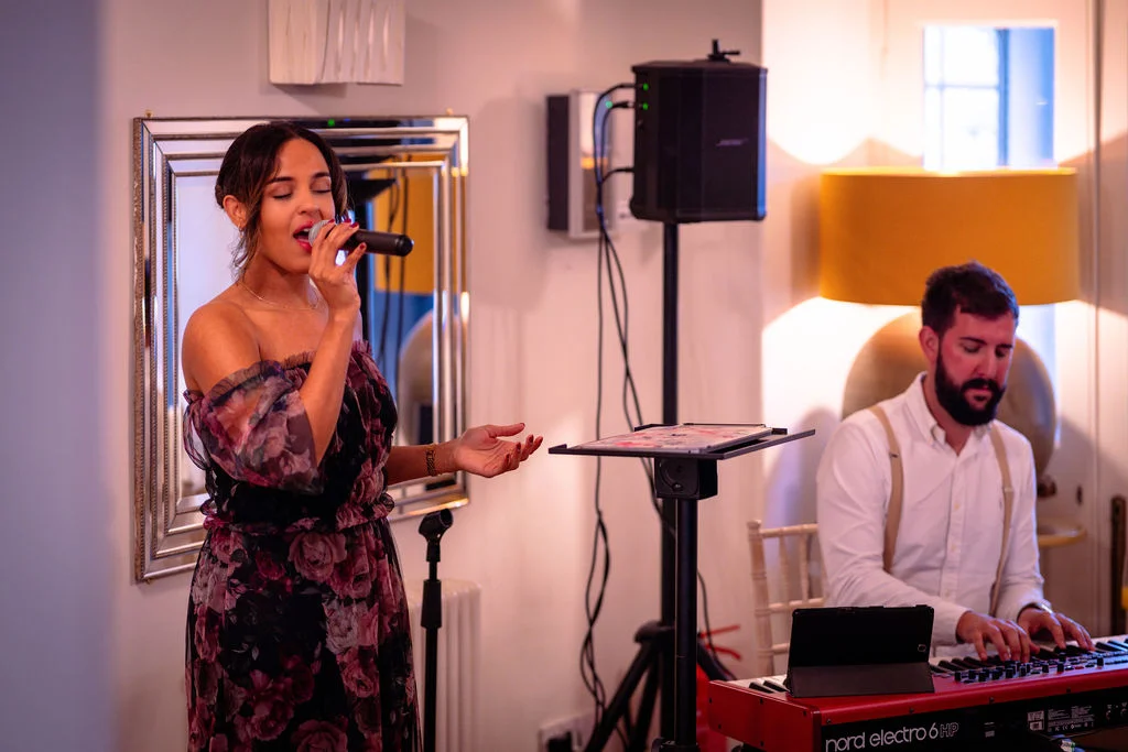 A live female singer in a floral off-the-shoulder dress performs with a microphone while a male musician plays a keyboard in the background.