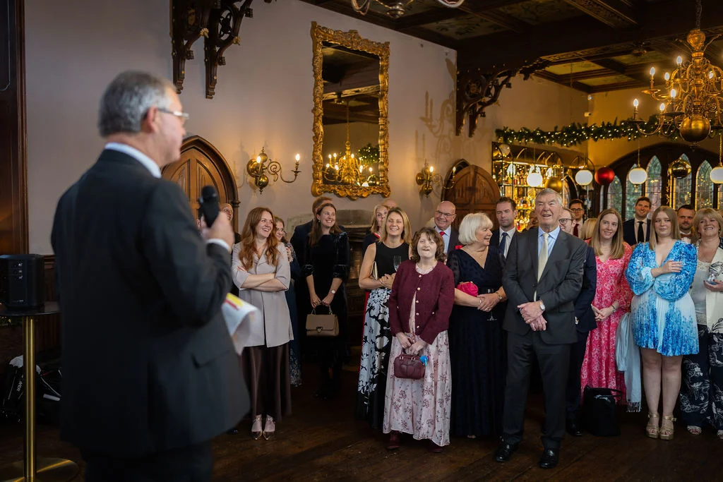 A wedding guest in a suit delivers a speech to an attentive audience of family and friends gathered in a grand panelled reception room.