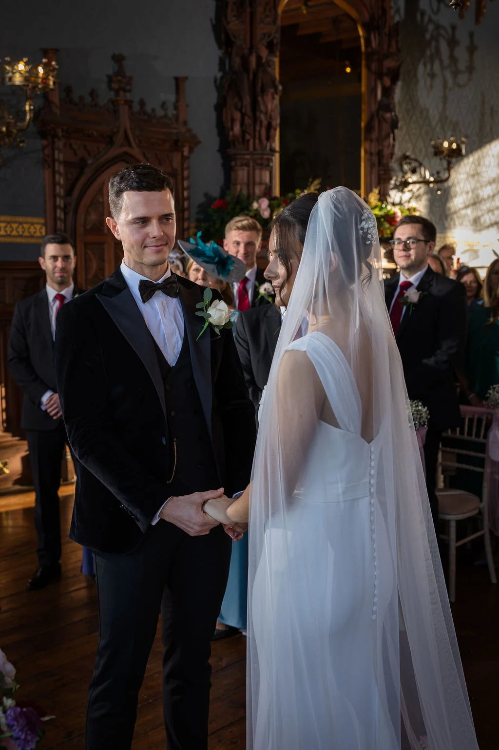 A groom in a velvet tuxedo and the bride in a sleek crepe gown hold hands at the altar during a traditional stately home wedding ceremony.
