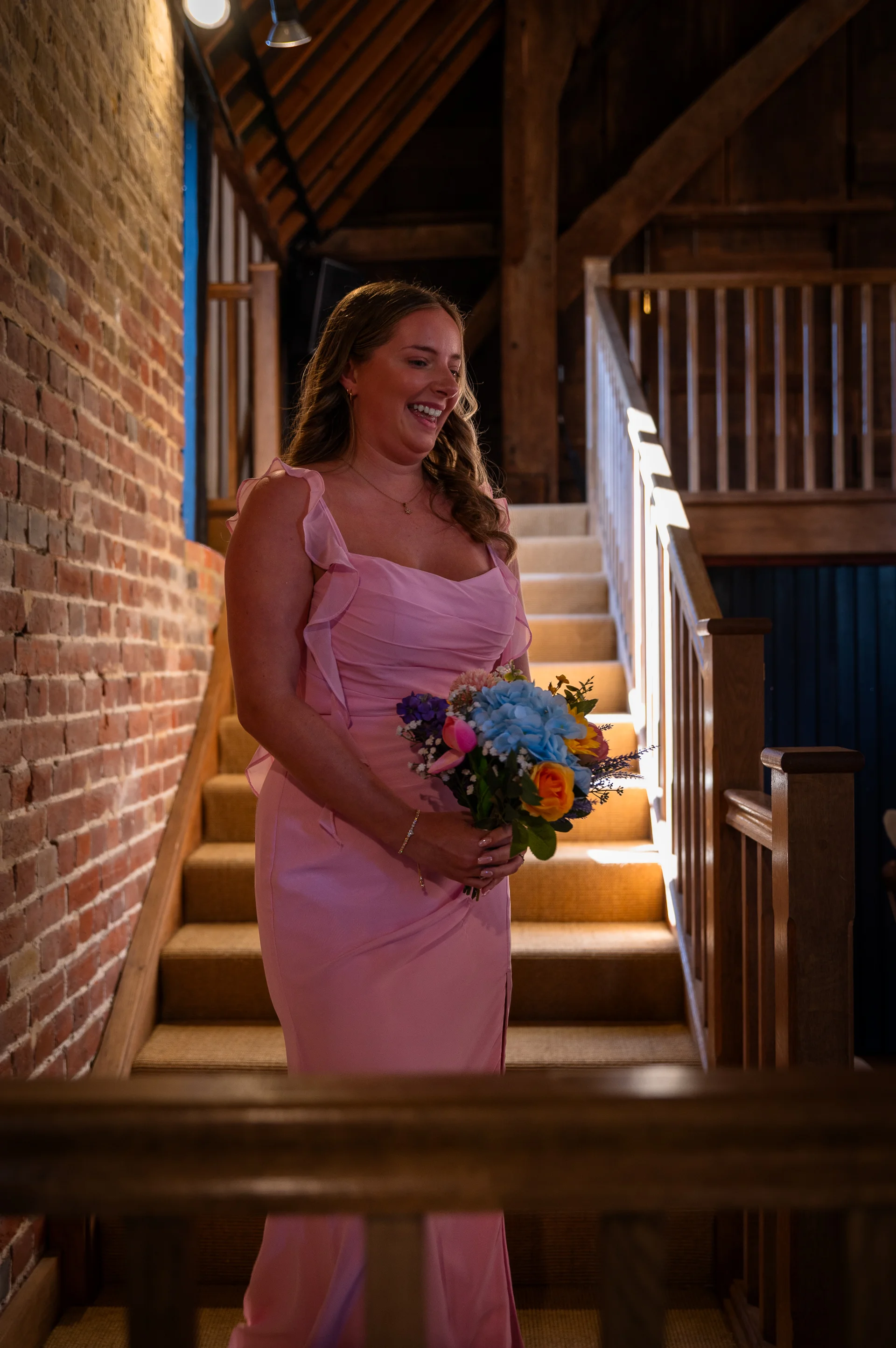 A smiling bridesmaid in a pink ruffled dress holds a colourful bouquet while standing on a wooden staircase inside an exposed brick barn.