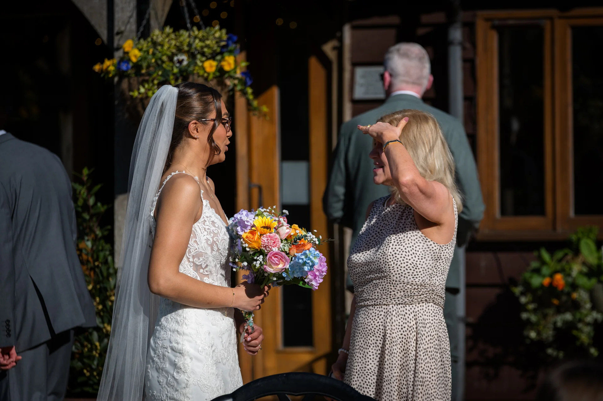 A bride in a lace gown and veil holds a colourful bouquet while chatting with a guest outdoors in bright sunlight.