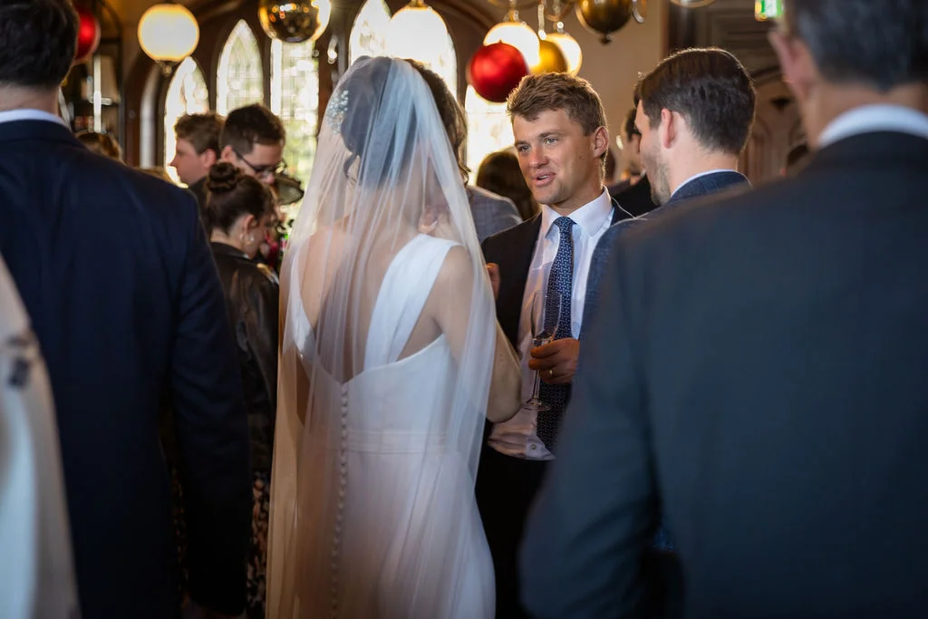 The newly married couple mingle with guests during a lively drinks reception, with the bride seen from behind wearing a veil over her gown.