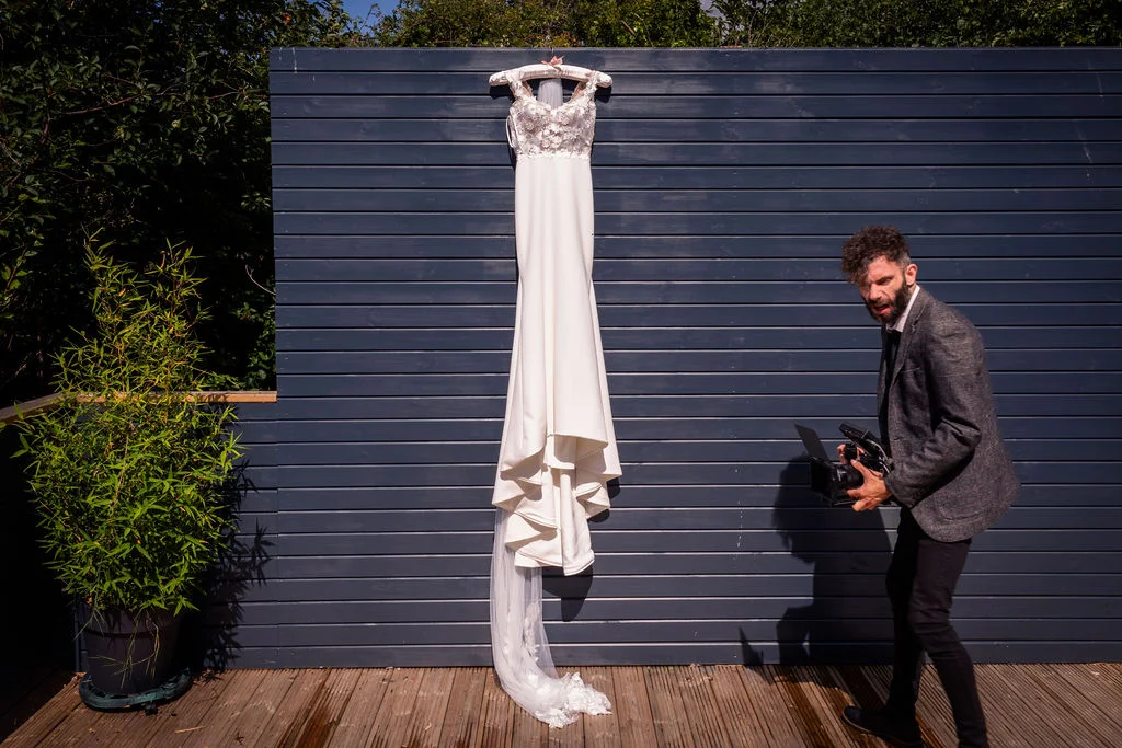 A videographer films a stunning crepe wedding dress with 3d lace detailing hanging against a dark wood panelled wall outdoors.