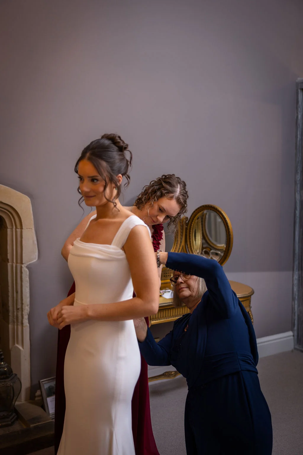 A mother and a bridesmaid help a radiant bride in a fitted white crepe gown get dressed inside a room during morning bridal preparations.