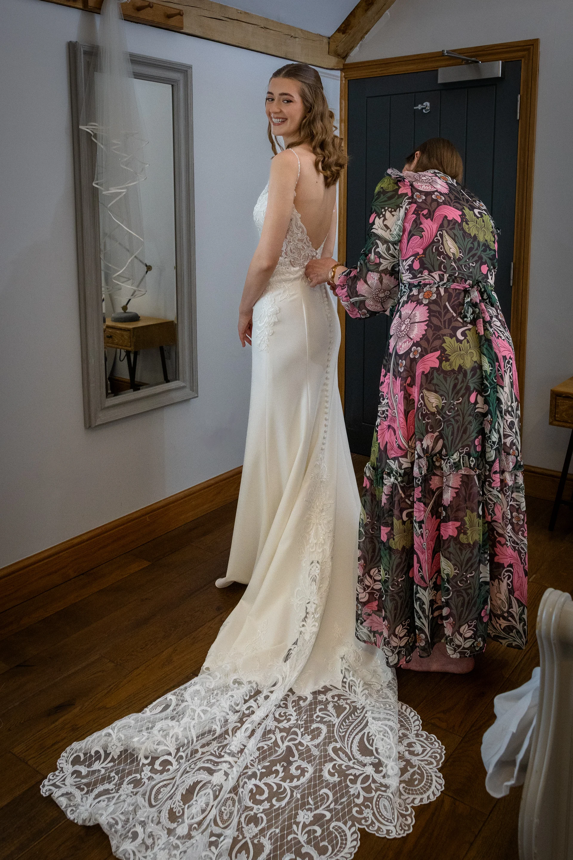 A smiling bride wearing an open-back lace and crepe wedding gown with a long train looks over her shoulder in a mirror while someone helps button the back of her dress.