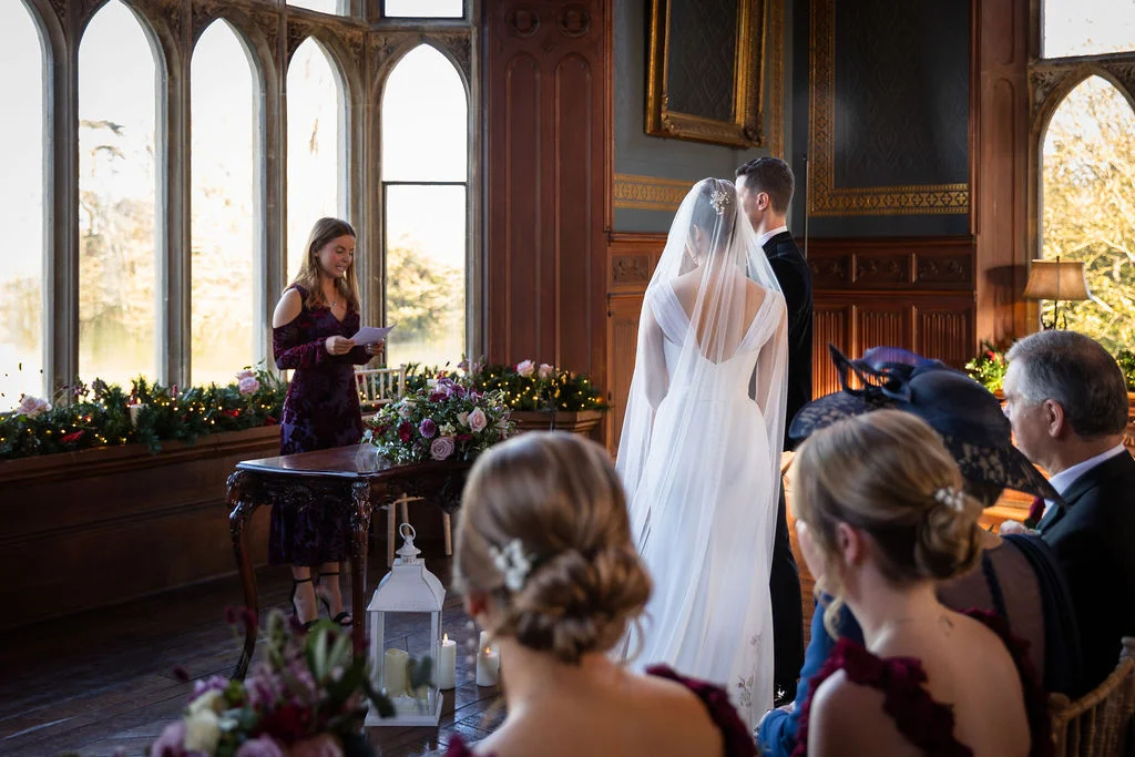 An intimate civil ceremony takes place inside a historic panelled room with large arched windows, where a couple stands facing a celebrant reading from a script.