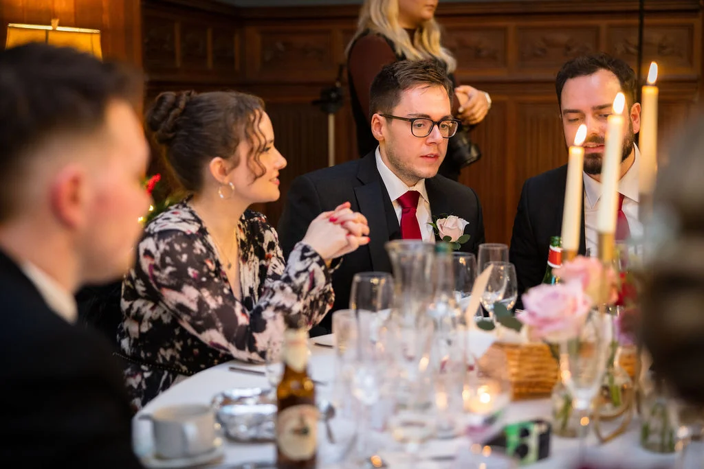 Wedding guests enjoy animated conversation and drinks at a candlelit banquet table inside a wood-panelled dining room during the wedding reception.