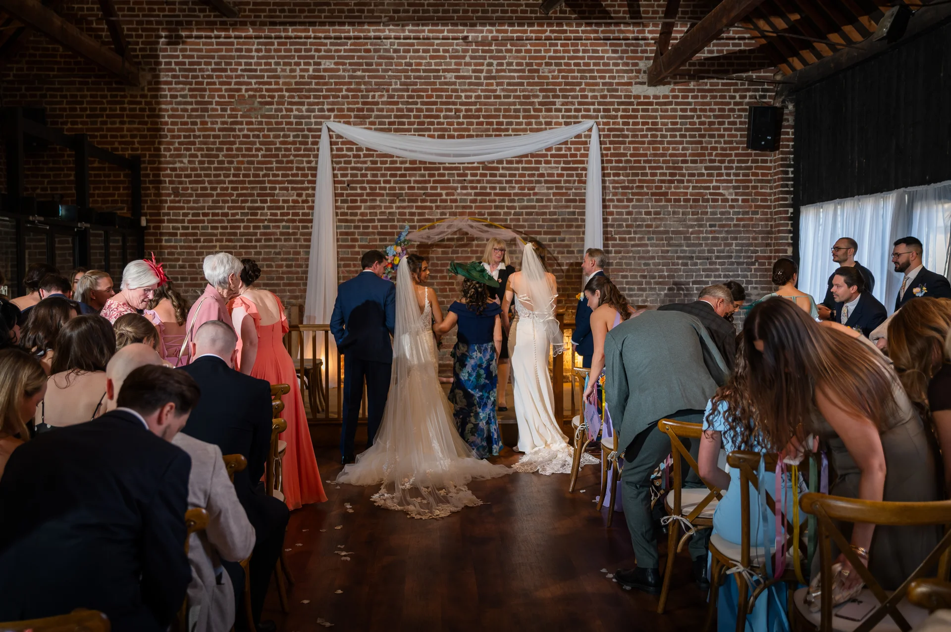 Guests are seated in a rustic brick barn venue as wedding participants walk down the aisle towards a draped ceremony backdrop.