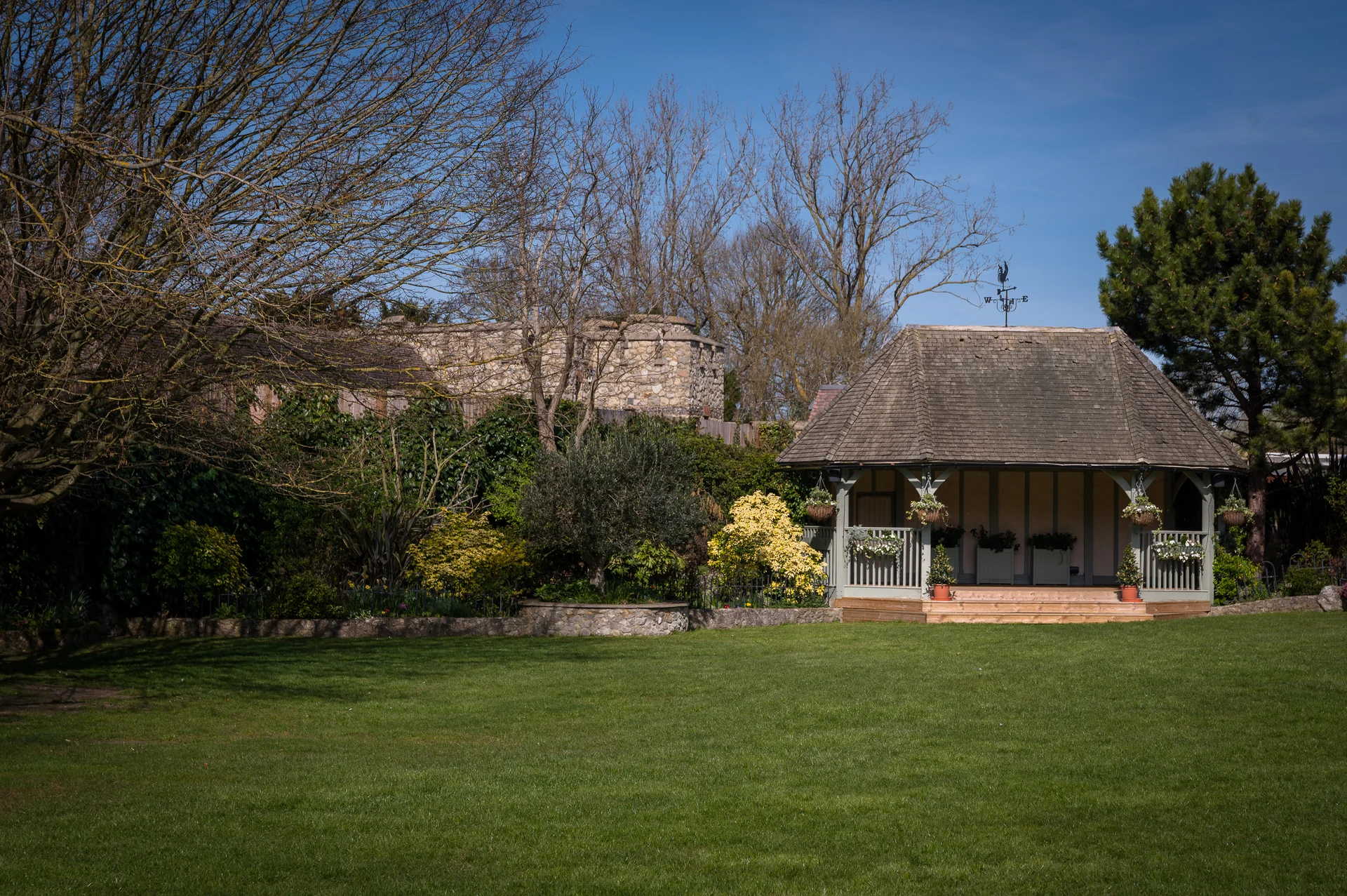 A picturesque wooden gazebo stands at the edge of a lush green lawn beneath a clear blue sky on a bright spring day.