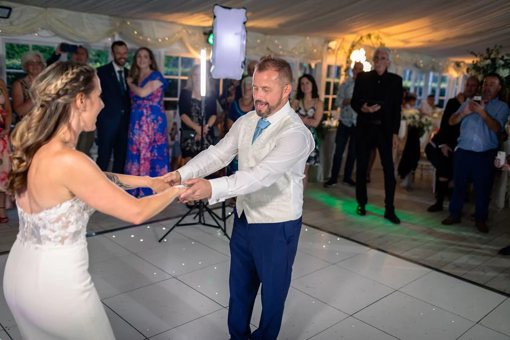 The bride and groom share an energetic first dance on a starlit dance floor inside a softly lit marquee surrounded by watching guests.
