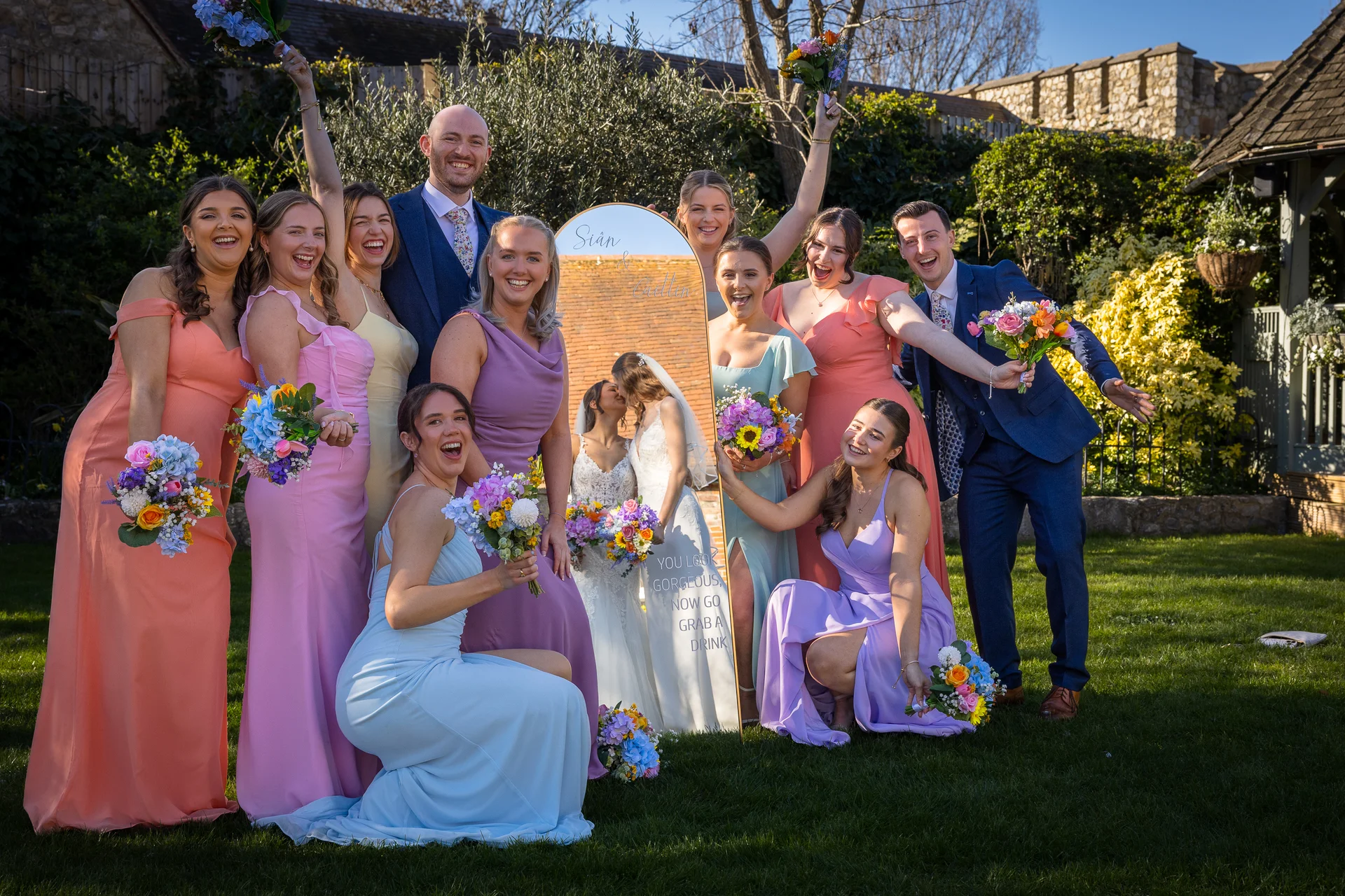 A joyful wedding party in pastel-coloured dresses and blue suits poses and laughs enthusiastically outdoors on a sunlit lawn next to a decorative mirror.