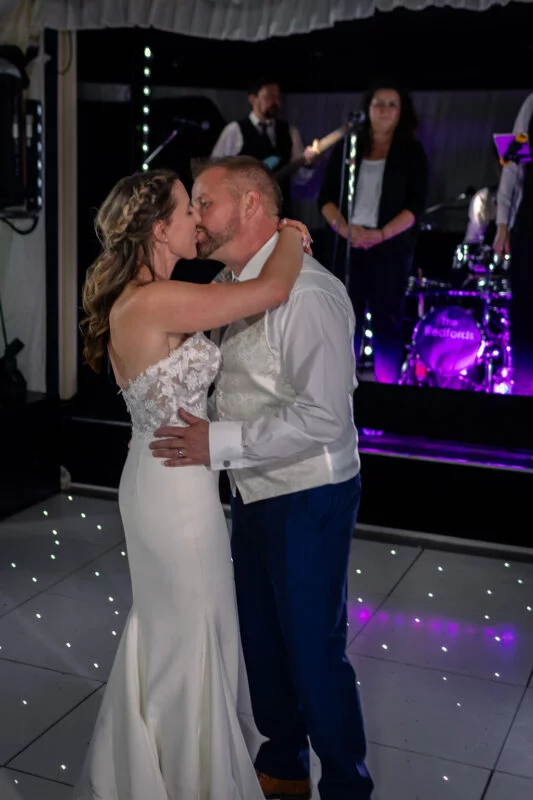 The newly married couple share a romantic kiss on an illuminated dance floor during their first dance, with a live wedding band performing on stage in the background.