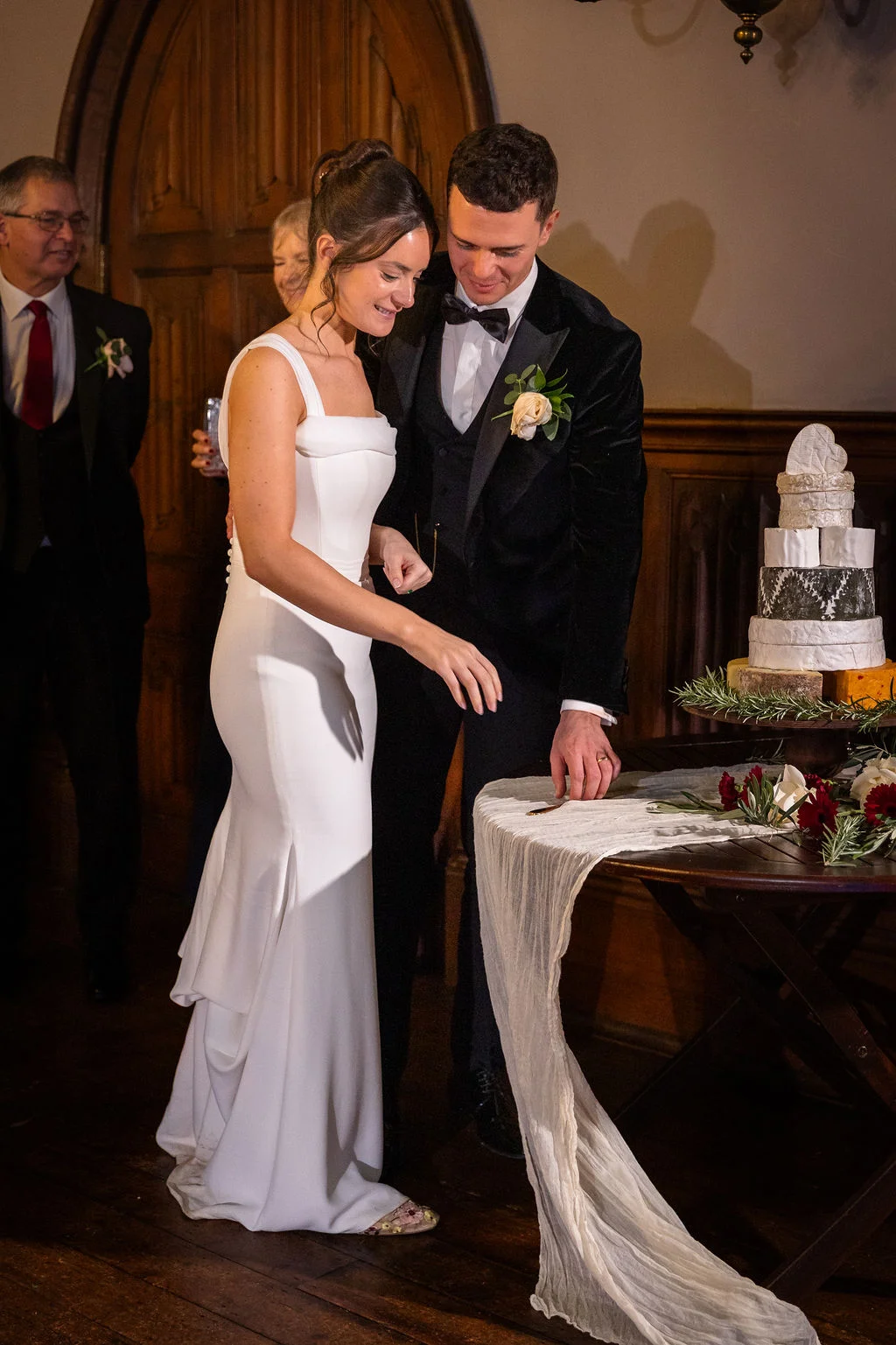 A smiling bride in a sleek crepe gown and groom in a velvet tuxedo prepare to cut a tiered cheese tower at their evening reception.