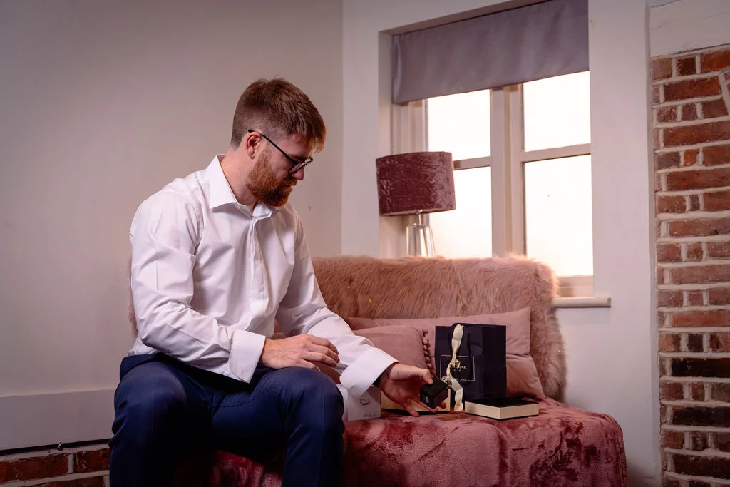 A groom in a crisp white shirt and blue trousers sits on a pink sofa near a window, examining a bottle of perfume during morning preparations.