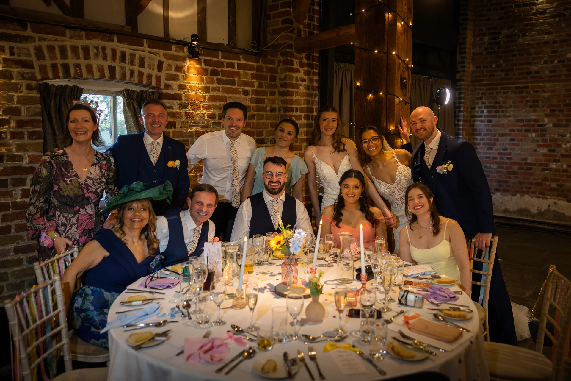 A large wedding party and guests smile and pose together around a candlelit round banquet table inside a rustic brick barn venue.