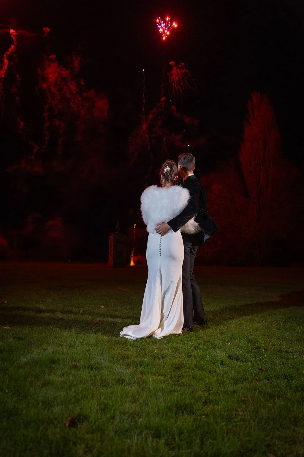 A newlywed couple stands on a lawn at night, embracing while watching a spectacular fireworks display light up the dark sky.