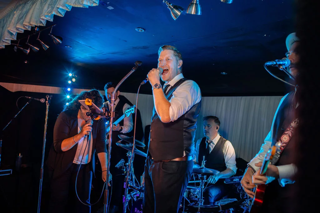 A live wedding band performs on stage under moody blue stage lighting, with the lead singer energetically singing into a microphone.