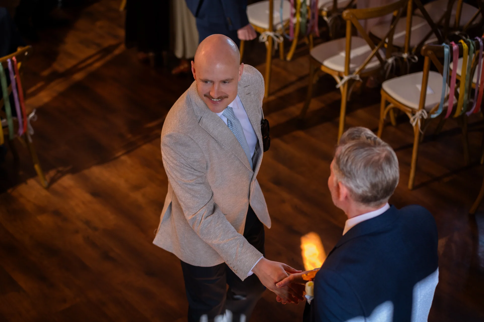 A high-angle shot captures a smiling groom in a light grey patterned suit shaking hands with another guest inside a wooden venue.
