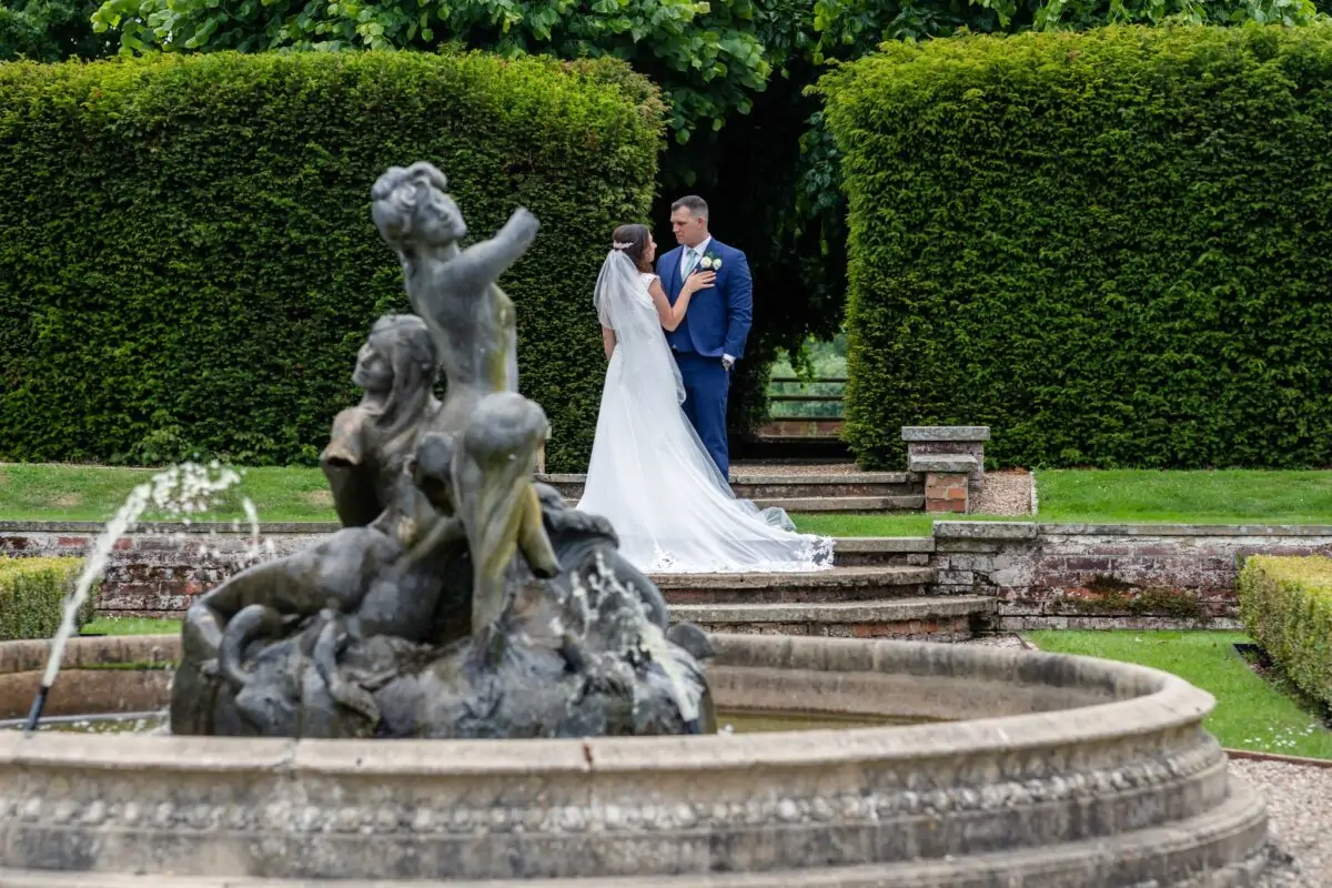 A newlywed couple stands together in a lush manicured garden near a stone fountain and topiary archways.